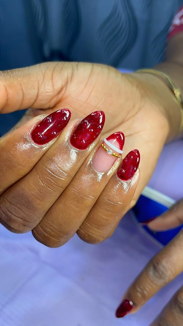 Deep red jelly almond nails with one nude accent nail framed in gold beads